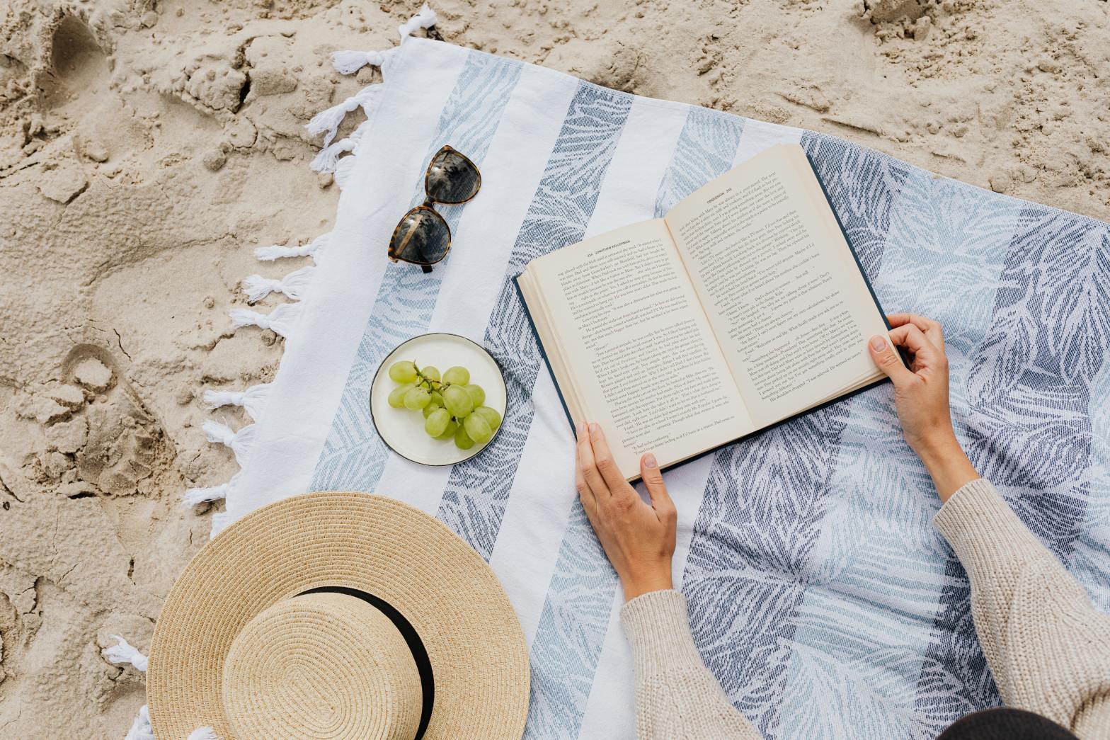 Sunglasses, A Plate of Grapes, A Hat, and Two Hands Holding a Book on a Beach Towel That is Laying on Sandy Beach